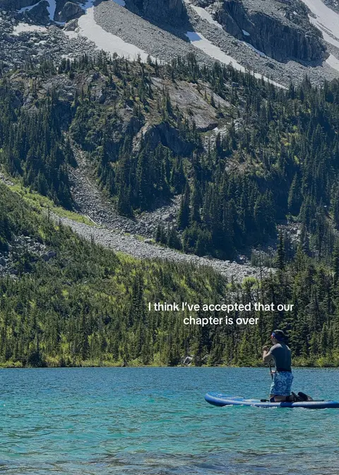 A scenic image of a serene mountain lake. A person kneels on a paddleboard, paddling towards the center of the lake. The water is a clear blue. A forested mountain backdrop rises above the lake. Text overlayed on the image reads: 'I think I've accepted that our chapter is over.'