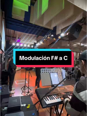 The cover image shows a live performance in a venue. A person is seen playing a keyboard, with a microphone stand and music stand in front of them. Other musicians and people are visible in the background, and there is stage lighting. Text overlayed in a rectangle on the image reads "Modulación F# a C".