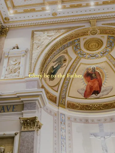 The image shows the interior of a decorated church. The ceiling is arched with detailed gold trim, and a painted scene with figures is visible. Text overlayed reads: "Glory be to the Father".