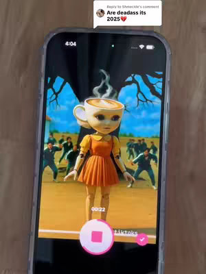 The cover image shows a phone screen displaying a scene from the TV series 'Squid Game', with a doll wearing a coffee cup as a hat overlaid on the image of the game's background, on top of a wooden text. The phone is set against a light brown wooden surface. The doll itself is dressed in an orange dress. At the top, there is a comment overlay that says: 'Are deadass its 2025❤️'.