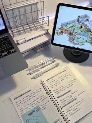 A clear, well-lit desk setup featuring studying materials. The foreground displays an open notebook with handwritten notes and sticky notes. Three pens lie beside it. A laptop is visible in the background on the left, and to the right, an iPad displaying a colorful map-like image. Various other desk accessories like a metal file holder and a white box are visible, suggesting a clean and organized workspace.