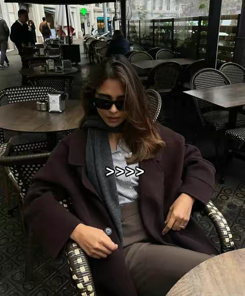 A woman wearing sunglasses and a dark coat sits in a cafe, the setting suggests a candid, posed shot with soft lighting. The background includes cafe tables and people.