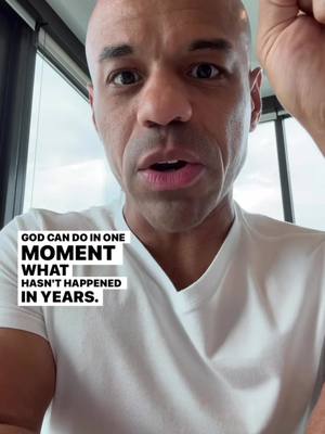 A medium shot features a bald man with a light skin tone, gazing directly at the camera with an open mouth. He is wearing a plain white crew-neck t-shirt. The background includes a window and an out-of-focus view of the sky. Overlayed text in a bold, black font reads: "GOD CAN DO IN ONE MOMENT WHAT HASN'T HAPPENED IN YEARS."