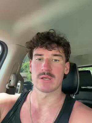 The cover image shows a man from the chest up inside a car. The man has short, dark, curly hair, a dark mustache, and a slight smirk on his face. He wears a black tank top and a silver chain. The interior of the car is visible on the left and behind the man. The shot looks like a selfie.