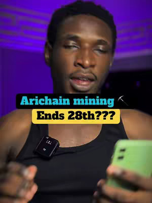A selfie-style video cover image featuring a dark-skinned man in a black tank top. He is looking at the camera, and a small microphone is clipped to his shirt. He holds a green smartphone in his right hand. The background has a purple hue. Overlaid on the image is a yellow and black text graphic that reads "Arichain mining Ends 28th???" with a pickaxe graphic.