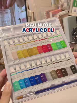 A close-up shot presents a tray filled with tubes of acrylic paint. The tray is white, and the paint tubes are aligned neatly. Each tube is labeled with text in Vietnamese. A paintbrush is placed along the bottom of the tray. The background is slightly blurred, featuring a glimpse of a laptop, some notebooks, and other stationery items. The lighting is soft and diffused, suggesting an indoor setting.