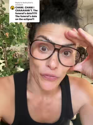 A woman with dark hair pulled back in a bun, wearing large black-framed glasses and a black tank top, is shown looking into the camera with a slightly distressed expression. She appears to be in an indoor setting with some greenery in the background. The image is a medium close-up, focusing on her face and shoulders.