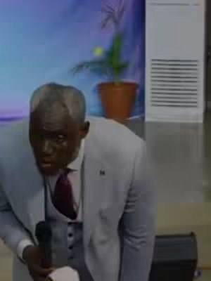 A man in a greyish-blue suit and burgundy tie is speaking into a microphone, possibly at a podium. The background is indistinct, with a glimpse of a potted plant and a large, light-colored structure that may be an air conditioning unit. The lighting appears to be indoor.