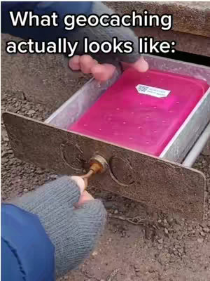 The image shows a person's hands, wearing grey gloves, interacting with what appears to be a geocache container. The container is a bright pink plastic box inside a metallic drawer or housing mechanism. The image is shot from a low angle, and the text overlay reads "What geocaching actually looks like:". The overall composition suggests a playful or humorous representation of the activity.