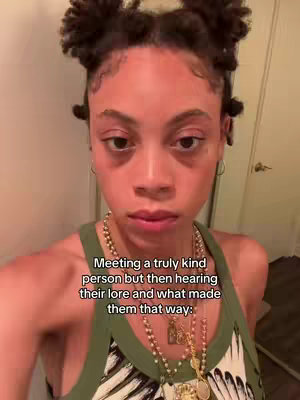 A selfie-style shot featuring a person with dark hair styled in two small buns on top of their head. They're looking directly at the camera with a subdued expression. They're wearing a green tank top and several necklaces. The background is a lightly-colored room with a partially visible door on the right side.
