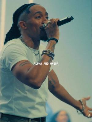 A man with dark hair and dreadlocks is singing into a microphone. He's wearing a white t-shirt and multiple bracelets and rings. The text \"ALPHA AND OMEGA\" is overlaid on the image.