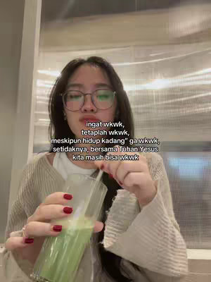 A young woman with dark hair and glasses is seen in a room, holding a glass with a green liquid in her hands. She is wearing a light-colored top. Text overlays are visible across the image.