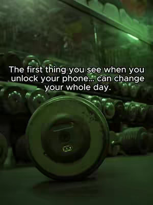 The image captures a gym setting with rows of barbells in the background. In the foreground, a weight plate is prominently displayed. Overlayed on the image is the quote: "The first thing you see when you unlock your phone... can change your whole day."