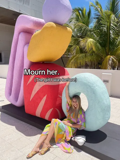 A woman in a colorful dress is seated in front of a large, abstract sculpture. The sculpture is comprised of multiple colorful, rounded shapes. The woman has one arm raised in the air and is smiling. Palm trees and a bright blue sky form the background.