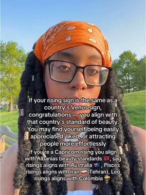 A selfie-style shot with a woman in the foreground. The woman is wearing glasses, an orange headscarf, and a nose ring. She has long, dark hair. Behind her is a blurred background with green trees and sunlight.