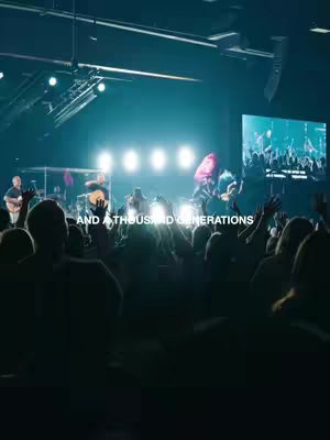 The image captures a worship concert scene. In the foreground, silhouetted figures of the audience are seen with raised hands, illuminated by stage lighting. The background features a stage with a band playing, and a large screen displaying additional images. The composition emphasizes a focus on the band and audience, with bright stage lights creating a dynamic and energetic atmosphere. Text overlays appear near the bottom of the image.