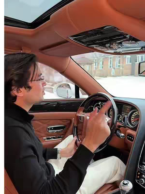 Inside a luxury car, a man in a black sweater and glasses is driving, pointing with his index finger. The car's interior is a warm beige, with a partially visible sunroof. The background shows buildings and a snowy environment.