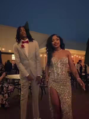A couple dressed in formal attire poses outdoors at dusk. The man, wearing a light-colored suit with a red bow tie, stands on the left with an arm at his side. The woman, in a sparkling gold gown with a high slit, is on the right.