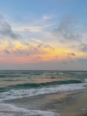 A breathtaking coastal scene at sunset. The sky blends hues of blue, yellow, and pink. The ocean transitions from a deep green to a lighter turquoise as it approaches the shore where the sand sits still. The image evokes a sense of tranquility and natural beauty.