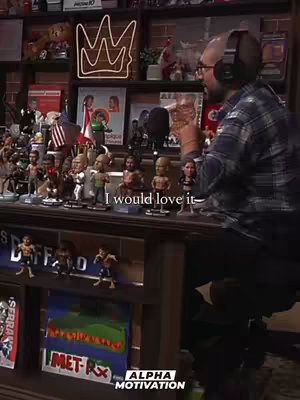 The cover image shows a man in a plaid shirt and headphones seated in front of a desk. Behind him on a shelf are various sports memorabilia figurines, awards, and what appears to be a neon crown. The man is holding a clear glass in one hand. The visible text on the screen says "I would love it-"