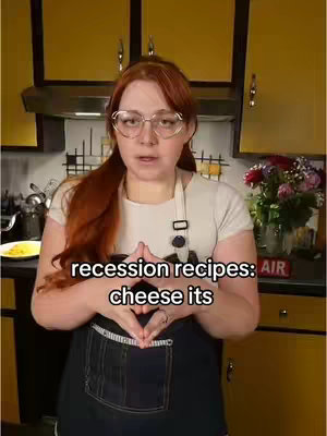 A woman with red hair and glasses is in a kitchen, wearing an apron and preparing a recipe. The text overlay says 'recession recipes: cheese its'. The kitchen has yellow cabinets and appears to be in a home setting, and there are some flowers on the counter.