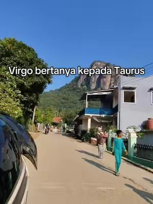 An outdoor shot of an asphalt road with a mountain in the background. A person in a green outfit is walking on the right side of the road, while a house and some people are seen to the right. The sky is blue, and the overall scene appears to be daytime