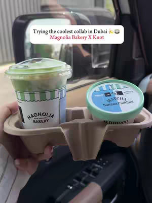 A hand holding a tray with two containers of food, inside a car. The tray has a Magnolia Bakery container on the left and a Matcha Banana Pudding container on the right. A text overlay floats above the containers. The background consists of the inside of a car and partially visible outdoor scenery.
