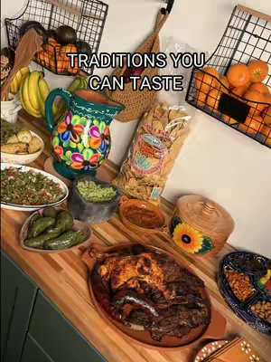 An overhead shot features a wooden surface laden with various Mexican food items. A large, colorful, ornate pitcher is to the left of center, next to a bowl of what appears to be guacamole. A platter of meat sits in the foreground with others foods in black wire baskets and bowls. The words 'TRADITIONS YOU CAN TASTE' are in a light font over the image.