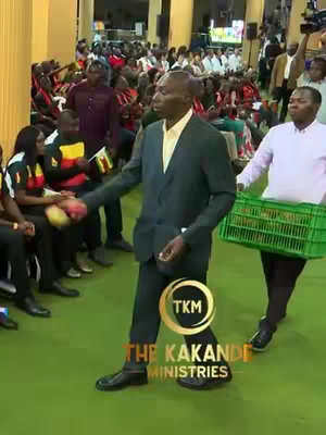 The image shows a man in a dark suit walking towards the viewer, shaking hands. A man to his right carries a green crate. The background is a room with many people. There is text overlayed on the image.