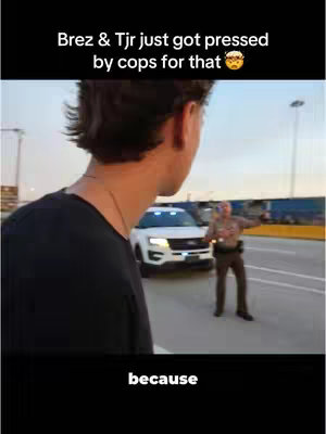 A person in a black shirt is seen from the back, looking towards a police car with flashing lights, along a highway. A police officer is gesturing to the person. Text overlays the image.