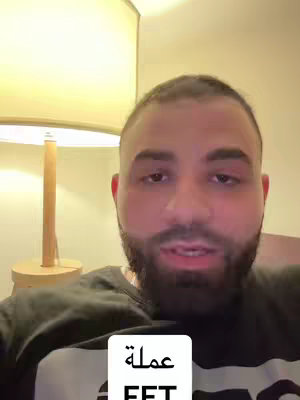 A man is in front of the camera with a lamp on the left and a sign at the bottom. The man has a beard. The background is beige.