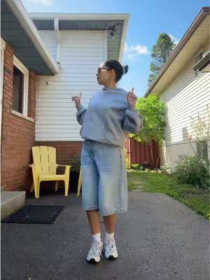 A woman stands outside in a sunny area, appearing to dance or celebrate with her arms up and fingers pointing towards the sky. The background shows houses and green spaces and suggests an outdoor setting.