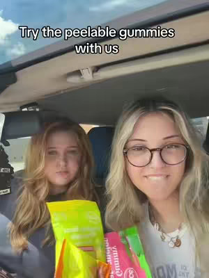Two young women are seated inside a vehicle, holding colorful bags of peelable gummies. The background is blurred, suggesting movement or an outdoor setting.