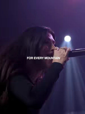 A person is singing into a microphone on stage, with stage lighting in the background. Text overlayed on the cover image reads, "FOR EVERY MOUNTAIN."