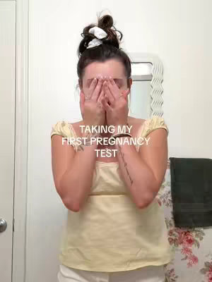 A woman stands in a bathroom with her hands covering her face. She is wearing a yellow top and white pants. The background includes a floral wallpaper and a decorative mirror. Text overlay indicates she is taking a pregnancy test.
