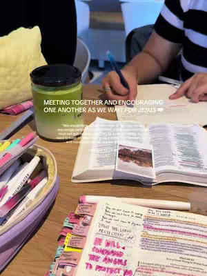 A close-up shot of a wooden table with an open Bible, a jar, and writing supplies. The Bible is open to a page with a picture in the center. A person's hand is visible, holding a blue pen and writing in a notebook. Additionally, there is a jar filled with a green substance, a pencil case and highlighters.