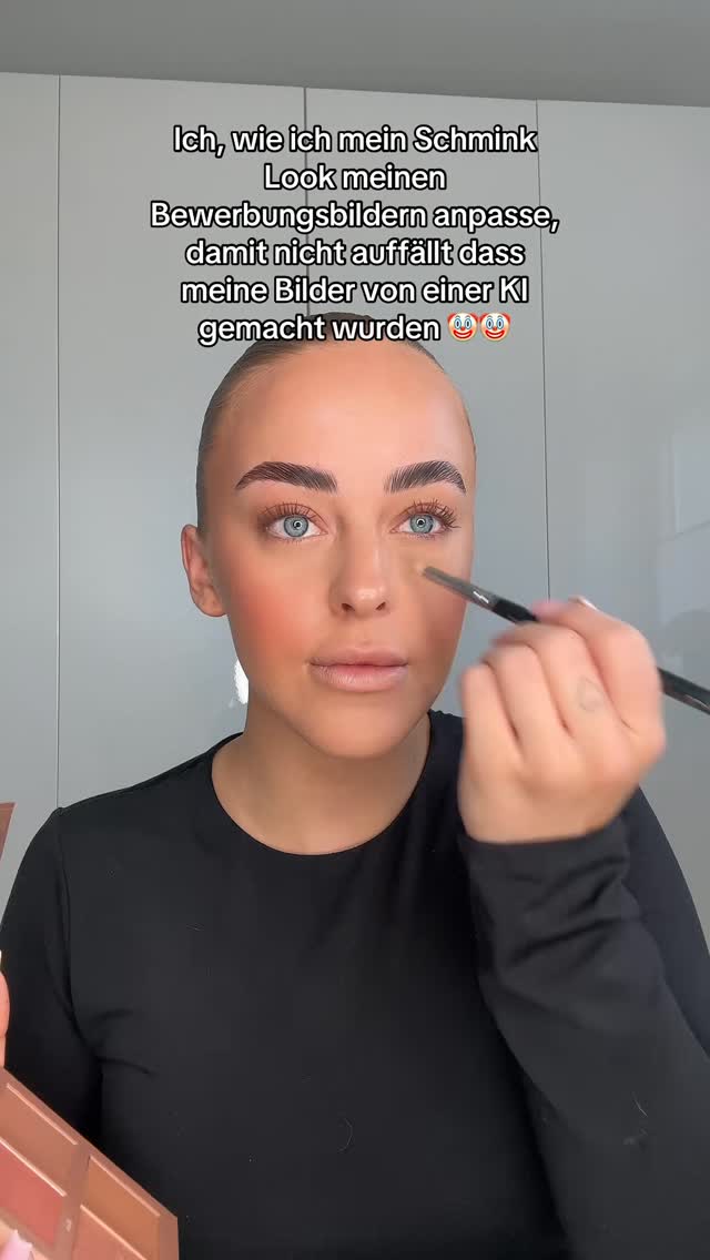 A selfie-style shot of a woman applying makeup. She is indoors, likely in front of a neutral-colored wall. The woman is fair-skinned with visible blue eyes, and she is holding a makeup brush near her eye. She is wearing a black long-sleeved shirt.