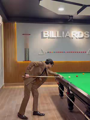 A man in a brown suit is positioned to take a shot at a billiards table. He is holding a pool cue and aiming at the cue ball. The background includes the word 'BILLIARDS' above a wall, the cue sticks, and colorful billiard balls. The lighting is well-lit, highlighting the scene and colors. The overall composition showcases a refined leisure activity.
