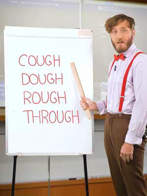 A man in a white shirt, red suspenders, and a bow tie stands to the left of a white board, pointing to the words written on it, with a light colored background.