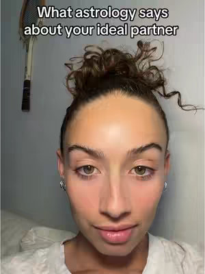 A selfie-style shot featuring a woman with brown curly hair in a bun, wearing a white top. Her face is the primary focus.