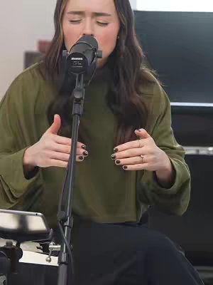 A woman stands in front of a microphone with her eyes closed, singing. She wears an olive-green long-sleeved top and black bottoms. Her hands are raised, with her fingers spread apart near her face. Her ringed fingers and dark nail polish are visible. The background includes a black piano.