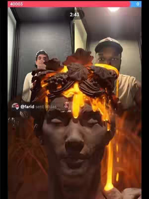 A selfie-style video with a dark, somewhat blurred background. In the foreground is a statue head sculpted in a classical style, with intricate details. The statue appears to be partially covered in a liquid that looks like molten gold, with the golden colored liquid dripping down its face. Behind the statue and the liquid, two dark-skinned men are visible. The overall tone of the image is stylized and artistic, combining contrasting elements.