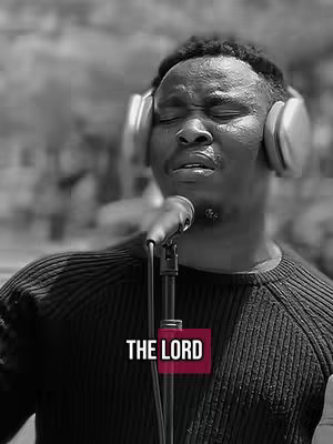 A black and white photo of a man with a shaved head wearing over-ear headphones and a black sweater, eyes closed, singing into a microphone. A text overlay that says "THE LORD" in a red box is placed near the bottom.
