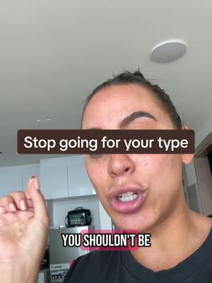 A woman with dark hair and visible freckles is the central figure. She's positioned from the chest up, wearing a black top. The background shows a brightly lit space with white cabinets, hinting at an interior setting. Overlayed on the image are two text boxes, one reading 'Stop going for your type' in a bold sans-serif font within a dark brown rectangle, and another below it stating 'YOU SHOULDN'T BE' in white sans-serif.