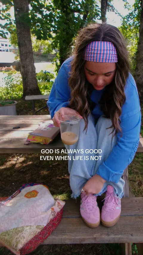 A young woman with long, wavy brown hair is sitting on a wooden picnic table outside with a bible, drink, and bag, wearing a blue shirt and headband. She has a neutral expression, and the sunlight creates natural shadows. The background features trees. The image is well-lit and the composition is centered.