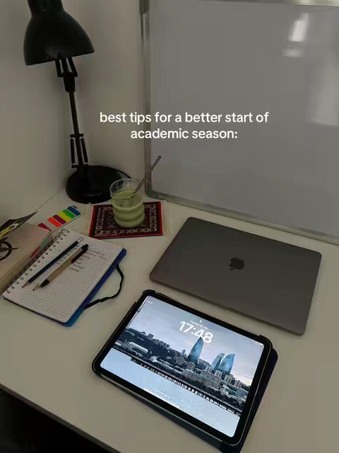 The image showcases a well-lit workspace arranged on a white desk. A black desk lamp and a piece of paper with colourful stickers are visible on the left. In the center, a green drink sits atop a decorative mat next to a notebook and pens. A closed laptop displays the Apple logo and an open iPad, showing an eye-catching city view, adds a modern touch. The overall scene is bright, clean, and inviting, hinting at an organized and productive study environment.