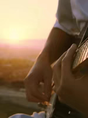 A close-up shot focuses on a person's arms and hands holding a guitar. The background is a soft, blurred view of a sunset, with warm colors. The scene is well-lit with natural light, creating a warm and inviting atmosphere.