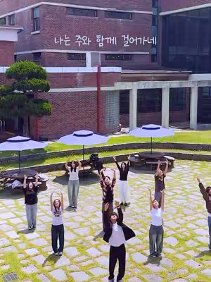 A wide shot shows a group of people raising their arms with buildings and umbrellas in the background. The scene is set outdoors, with a green lawn and brick buildings visible. Several people are visible, with their arms raised toward the sky. They appear to be in the middle of a performance or activity. The background features a brick building and several white umbrellas that are spread out. The overall impression is of a coordinated group activity taking place in a park-like setting.