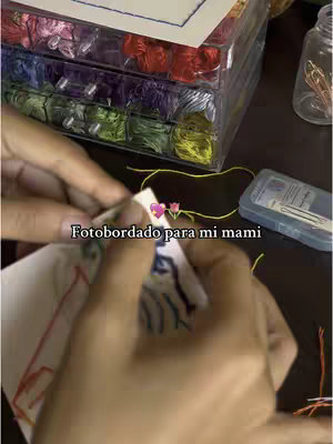 A close-up shot of hands embroidering a piece of cloth, with various colored threads, a plastic container with thread, and other craft supplies visible on a dark wooden surface. The text overlay says 'Fotobordado para mi mami'.