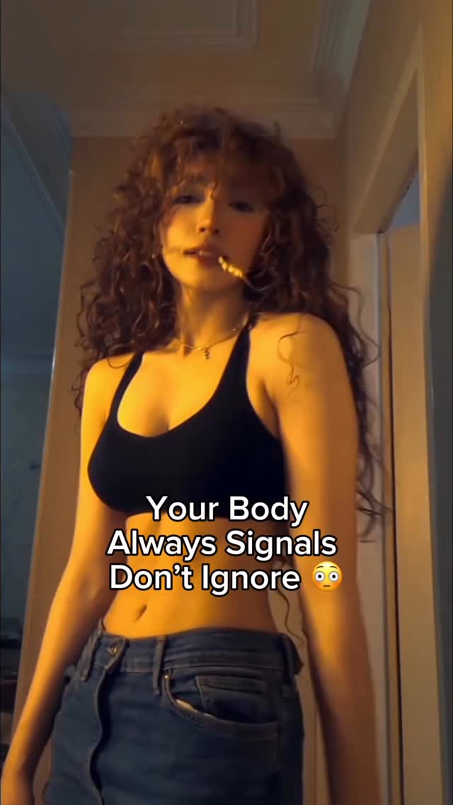 The image shows a woman with curly hair wearing a black top and jeans, standing in a room with warm lighting. Text overlayed on the image says, 'Your Body Always Signals Don't Ignore 😳'.